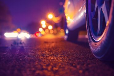 Night country road with glowing lanterns. Headlights of approaching cars. Close up view from the level of a parked car wheels.