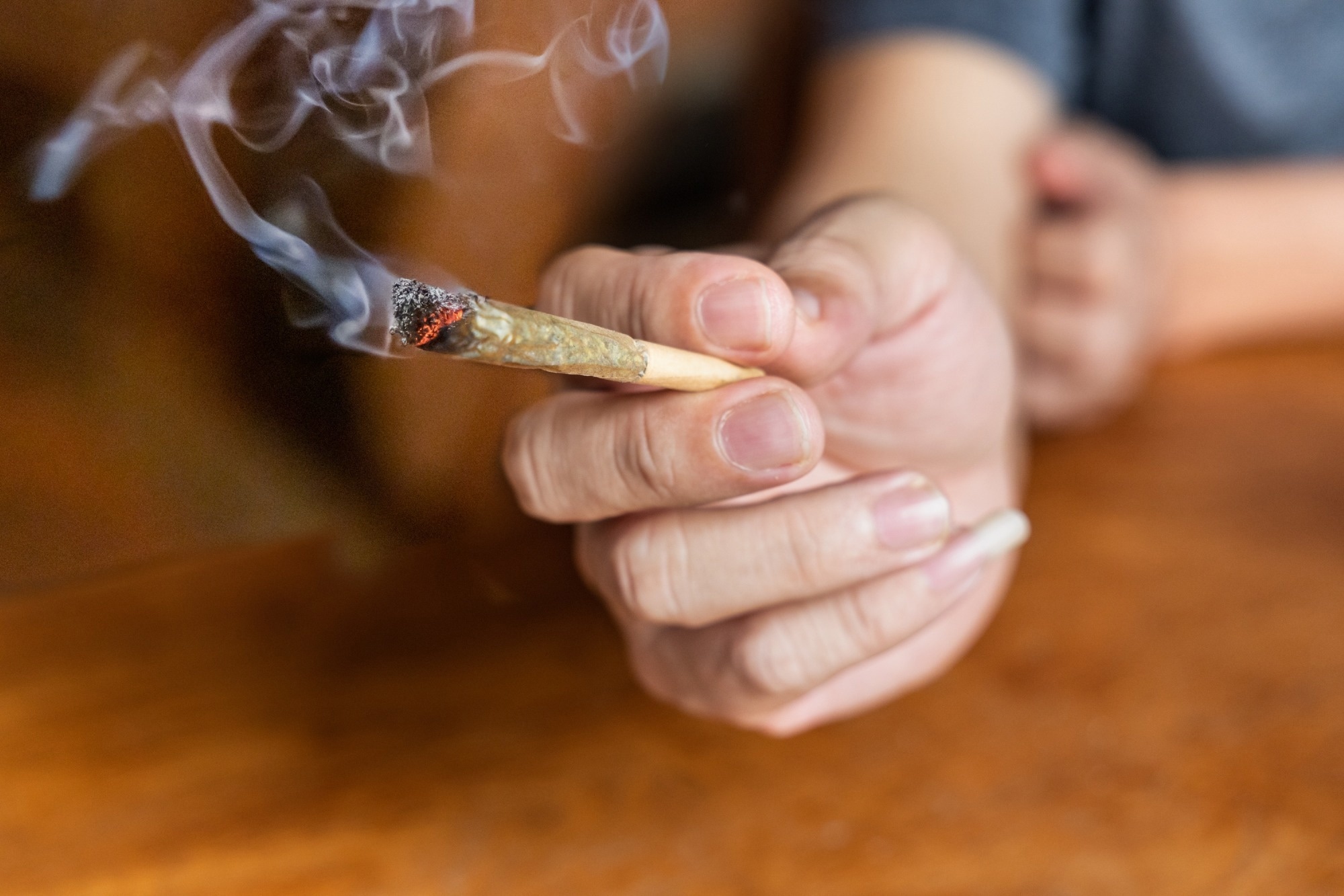 Finger holding lighted cannabis cigarette with smoke in dark room.