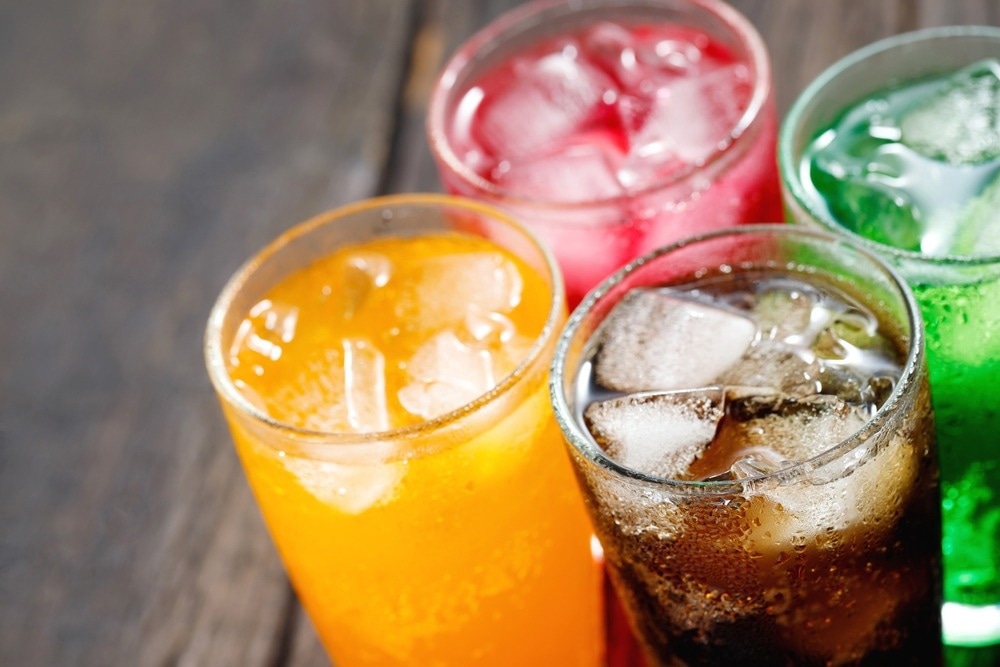 Four glasses of colorful sugar-sweetened beverages, including orange, red, green, and cola drinks, served over ice on a wooden table.