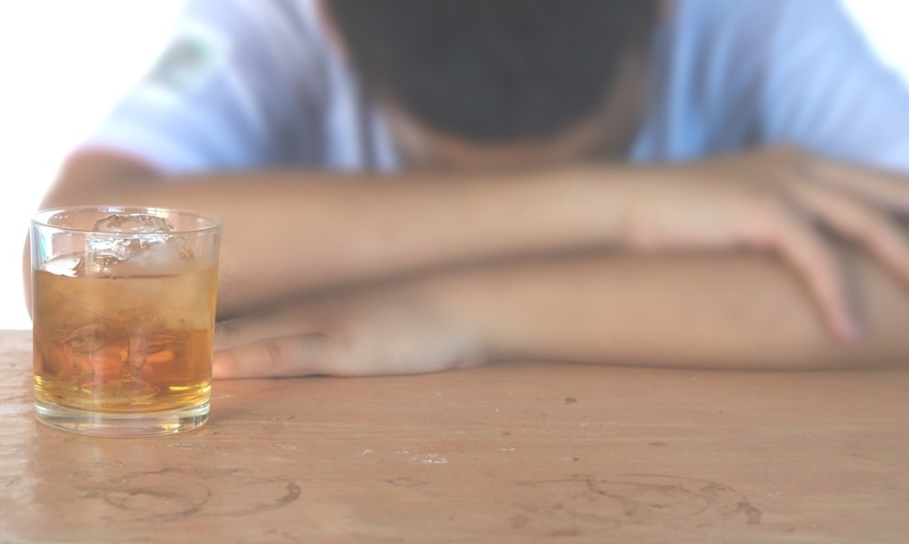 person with their head down on a wooden table, appearing fatigued or distressed, with a glass of whiskey on ice in the foreground. The image is blurred except for the drink, emphasizing alcohol consumption.