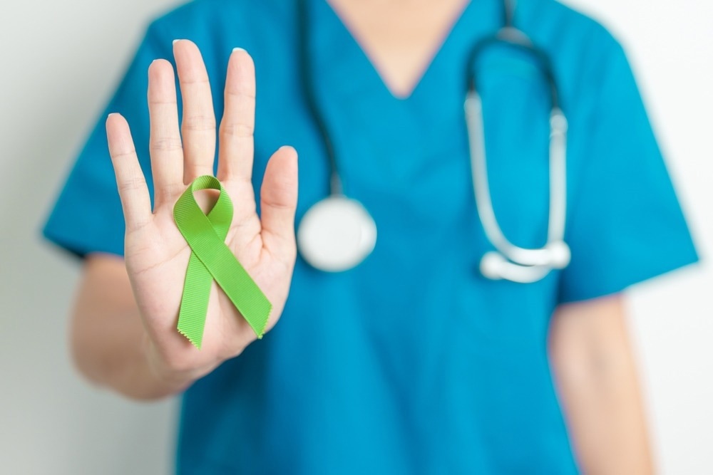 A healthcare professional wearing blue scrubs and a stethoscope holds up their hand displaying a green awareness ribbon, symbolizing support for Non-Hodgkin